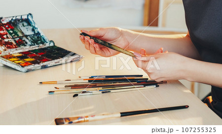 Art tools. Painting accessories. Unrecognizable woman artist hands with paintbrushes watercolor paint palette at artwork table studio workplace. Art tools. Painting accessories. Unrecognizable woman artist hands with paintbrushes watercolor paint palette at artwork table studio workplace. 107255325