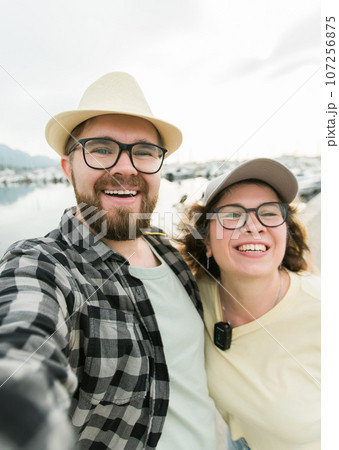 Millennial couple taking a self portrait laughing as they pose close together for camera on their smartphone outdoors in summer port marina with boats and yachts - travel and videocall and blog 107256875