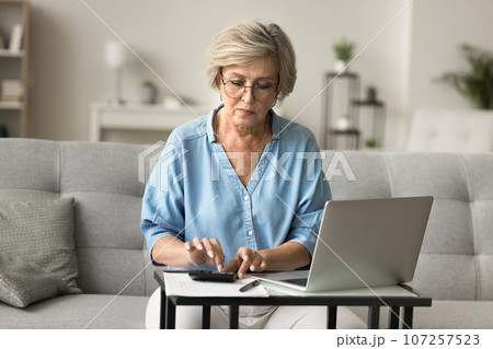 Serious older mature woman counting savings on calculator at laptop Serious older mature woman counting savings on calculator at laptop 107257523