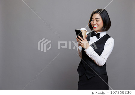 Smiling asian woman wearing receptionist uniform texting and scrolling social media while drinking coffee to go. Restaurant cheerful waitress enjoying tea in paper mug and using mobile phone Smiling asian woman wearing receptionist uniform texting and scrolling social media while drinking coffee to go. Restaurant cheerful waitress enjoying tea in paper mug and using mobile phone 107259830