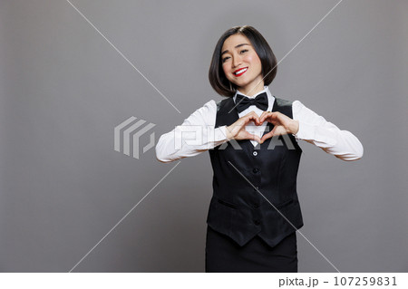 Cheerful smiling asian woman wearing waitress uniform showing heart symbol with fingers portrait. Happy receptionist showcasing love and affection gesture and looking at camera Cheerful smiling asian woman wearing waitress uniform showing heart symbol with fingers portrait. Happy receptionist showcasing love and affection gesture and looking at camera 107259831