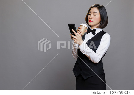 Asian waitress browsing social media using smartphone during coffee break. Serious woman receptionist wearing uniform texting on mobile phone and holding drink to go paper cup Asian waitress browsing social media using smartphone during coffee break. Serious woman receptionist wearing uniform texting on mobile phone and holding drink to go paper cup 107259838