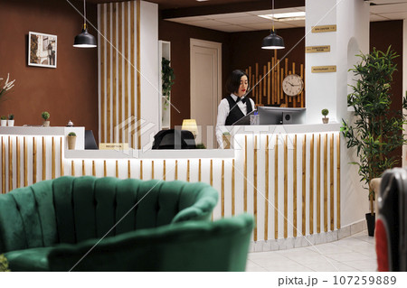 Hotel concierge writing booking form at front desk, preparing to help guests with accommodation at arrival. Female resort employee checking registrations in record, reception area. Hotel concierge writing booking form at front desk, preparing to help guests with accommodation at arrival. Female resort employee checking registrations in record, reception area. 107259889