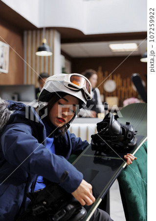 Activity-focused traveler adjusts wintersports gear in hotel lobby preparing for winter holiday at ski resort. Close-up of guest holding and checking snowboard. Excitement for snowy adventure. 107259922