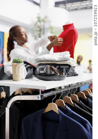Jackets hanging on rack and accessories on shelf in clothing store while assistant dressing mannequin on blurred background. Male blazers and belts in department shopping mall 107259923