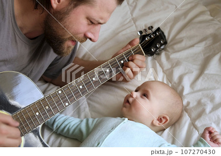 Bearded Father Playing Guitar to his Little Child. Baby Looking at Him, Listening and Smiling. Portrait of Happy Family at Home. Joyful Leisure. Handsome Caucasian Father and Cute Baby Boy Having Fun. 107270430