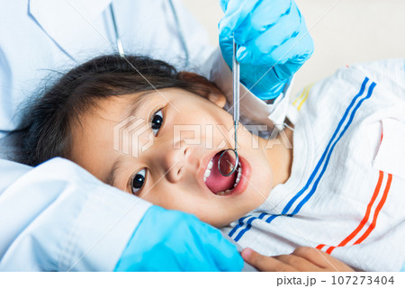 Dental kid health examination. Doctor examines oral cavity of little child uses mouth mirror to checking teeth cavity, Asian dentist making examination procedure for smiling cute little girl 107273404