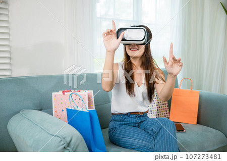 Asian young woman with shopping bags around wearing modern virtual reality headsets having experience in shopping online in living room at home, Happy female with VR headset on sofa 107273481