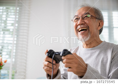 Asian senior old man enjoying holding joystick playing video game at home in living room, mature man hands using game controller, Funny on retirement elderly smile sitting on sofa life gaming 107273545