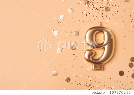 Gold candle in the form of number nine on peach background with confetti. Gold candle in the form of number nine on peach background with confetti. 107276219