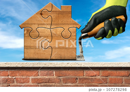 Gloved Hand Holding a Trowel and a Small Wooden Model House 107278298