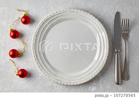 Empty white plate, red Christmas balls and cutlery on a gray stone table. New Year's menu 107278565