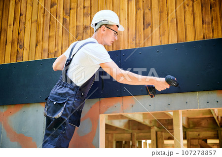 Carpenter constructing wooden framed house. Man worker in glasses cladding facade of house with cement particle boards, fastening them with screwdrivers. Concept of modern eco-friendly construction. 107278857