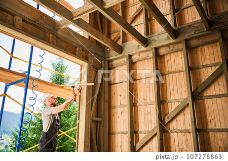 Carpenter constructing wooden frame, two-story house near forest. Bearded man hammering nails with hammer while wearing protective helmet and work coveralls. Concept of modern ecological construction. 107278863