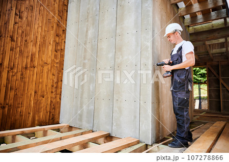 Carpenter constructing wooden framed house. Man worker cladding facade of house with cement particle boards, fastening them with screwdriver. Concept of modern eco-friendly construction. 107278866