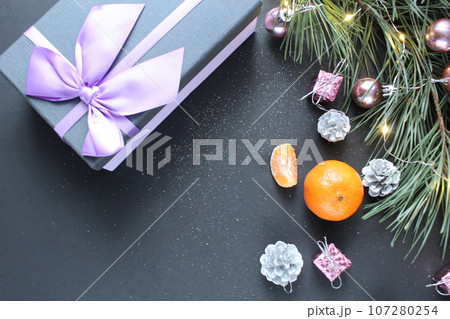 still life New Year's Christmas. A gift with a bow lies on a black background next to a branch of spruce pine trees mandarin toys Christmas tree with a place for text copyspace top view still life New Year's Christmas. A gift with a bow lies on a black background next to a branch of spruce pine trees mandarin toys Christmas tree with a place for text copyspace top view 107280254
