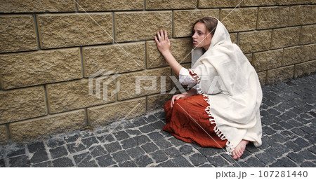A girl in poor clothes sits on the ground 107281440
