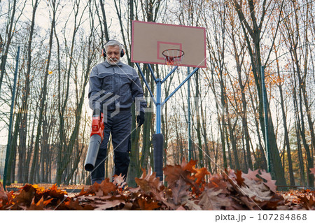 Aged janitor taking care of park area during autumn season. Low angle view of bearded man in overalls using electric blower to remove dry leaves at outdoors basketball court. Seasonal work concept. 107284868
