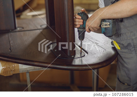 Close up of carpenter fixing table with electric drill standing in carpentry workshop and looks away Close up of carpenter fixing table with electric drill standing in carpentry workshop and looks away 107286015