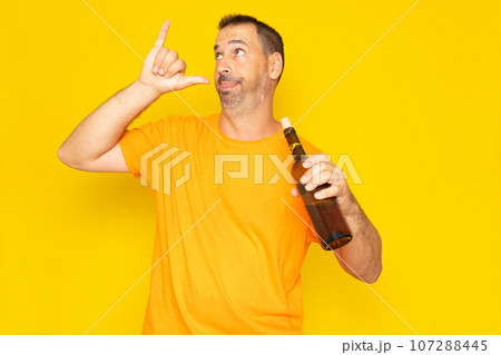 Hispanic man with beard in his 40s making a go drunk gesture with a bottle of wine in his hand, isolated on yellow studio background. 107288445