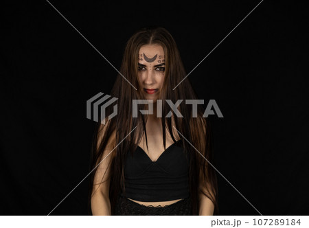For halloween, a female witch stands in the dark with tousled hair falling over her face. Portrait of a Vedbma with gothic symbols on her face and long black hair. Witch concept for halloween. 107289184
