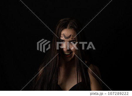 Evil laughing at the top of her voice, the witch looks into the camera on a black background in the twilight. On Halloween, a female witch grinning in a terrible grin looks at the camera. 107289222