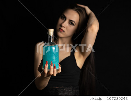 For the holiday of halloween, a gothic witch woman holds out a bottle with a potion on a black background. The witch holds in her hand a bottle of luminous liquid. Halloween concept. 107289248