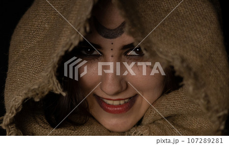 A grinning witch peeks out from under the hood at Halloween night. A beautiful female witch with a hood thrown over her head smirks while looking at the vamer on a black background. Close-up. 107289281