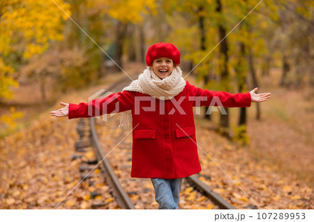 Caucasian girl in a red coat and beret walks along the railway tracks in the park in autumn. Caucasian girl in a red coat and beret walks along the railway tracks in the park in autumn. 107289953