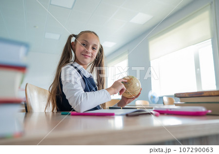 Caucasian schoolgirl sits at her desk at school and studies the globe.  107290063