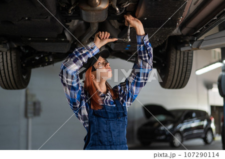 Female mechanic unscrew the nuts on the bottom of the car that is on the lift. A girl at a man's work. 107290114