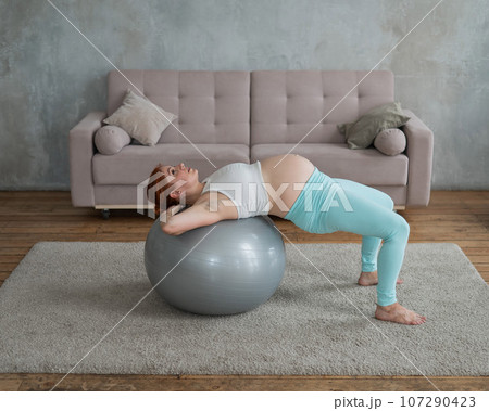 Pregnant red-haired woman doing exercises on fitness ball at home. Pregnant red-haired woman doing exercises on fitness ball at home. 107290423