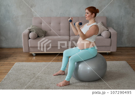 Pregnant woman doing exercises with dumbbells while sitting on a fitness ball at home. Pregnant woman doing exercises with dumbbells while sitting on a fitness ball at home. 107290495