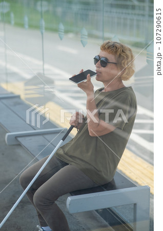 Elderly blind woman sitting at a bus stop and using a smartphone.  107290615