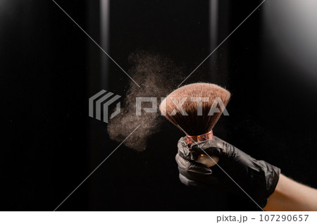 A woman shakes a large powder brush with clouds of dust on a black background. Treatment after instant tanning 107290657