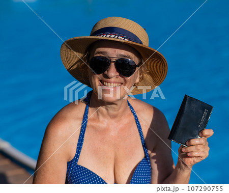 An elderly woman in a hat and sunglasses holds a passport while sitting by the pool. Vacation in retirement. An elderly woman in a hat and sunglasses holds a passport while sitting by the pool. Vacation in retirement. 107290755