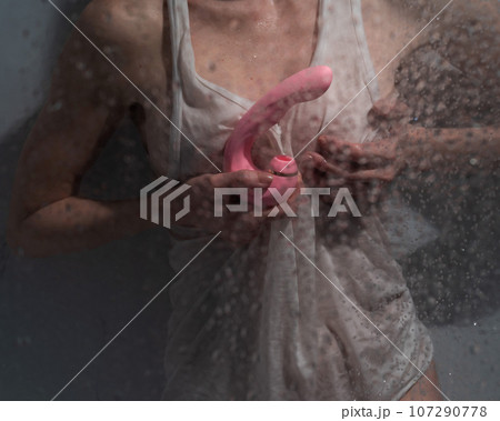 A woman in a white tank top stands in the shower and holds a curved pink sex toy.  107290778