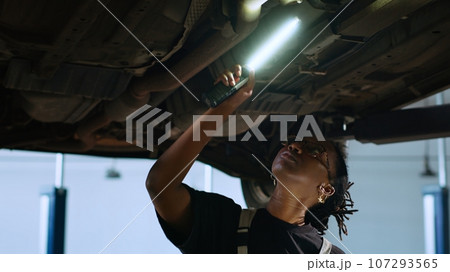 Licensed mechanic standing underneath suspended car in garage workspace, using work light to check for damages and tablet to order new replacing parts after finding issues during annual maintenance Licensed mechanic standing underneath suspended car in garage workspace, using work light to check for damages and tablet to order new replacing parts after finding issues during annual maintenance 107293565