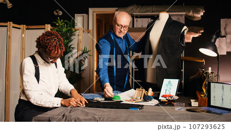 Couturier and trainee creating clothes in atelier, working with tailoring tools for manufacturing process. Apprentice learning handcraft skills from experienced senior tailor. Handheld shot. Couturier and trainee creating clothes in atelier, working with tailoring tools for manufacturing process. Apprentice learning handcraft skills from experienced senior tailor. Handheld shot. 107293625