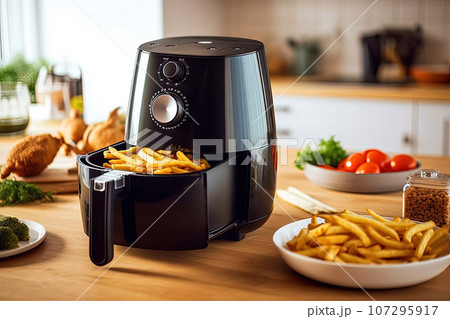 An air fryer in black color or an oil-free frying machine placed on a wooden table in a contemporary kitchen with plates of fried potatoes and tomatoes 107295917