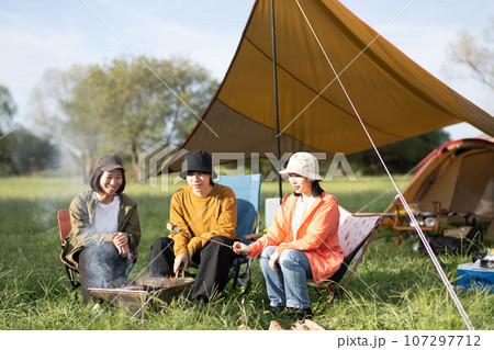 キャンプを楽しむ若い男女・焚き火・焼きマシュマロを焦がして笑う3人・アウトドアイメージ 107297712
