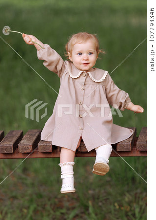 A little girl in a dress sits on a wooden bench in the park 107298346