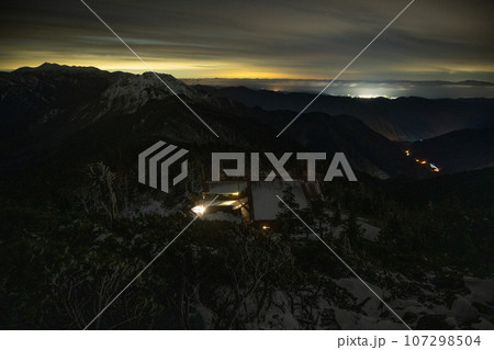 11月の曇天の下、北アルプスの稜線から山小屋の灯りと岐阜県高山市の夜景 107298504