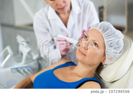 Young female cosmetologist doing microcurrent procedure to adult woman 107298506