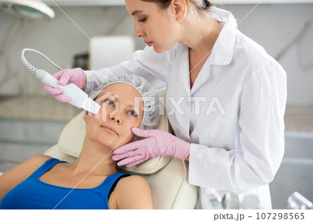 Young beautician doing procedure for face lifting and Soften skin keratin, deep clear skin dirt of adult woman laying in salon. Hardware cosmetology Young beautician doing procedure for face lifting and Soften skin keratin, deep clear skin dirt of adult woman laying in salon. Hardware cosmetology 107298565