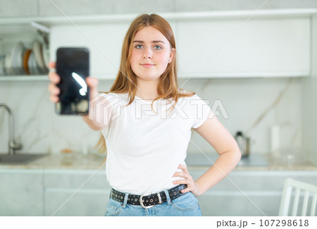 Smiling teenage girl presenting smartphone with blank black screen with copy space in modern cuisine. Free copy space for your design or advertisement Smiling teenage girl presenting smartphone with blank black screen with copy space in modern cuisine. Free copy space for your design or advertisement 107298618