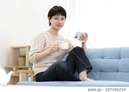 ソファーに座りスマートフォンを使う若い女性 107300224