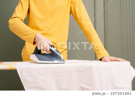 Thin human in yellow longsleeve standing, ironing white shirt on pressboard using electric flatiron. Man do housework. 107301981