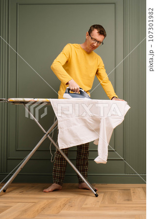 Mature man in glasses ironing snow-white shirt on pressboard using flatiron. Soigne tall human working around house. 107301982