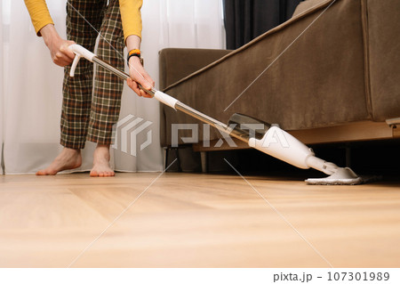 Unrecognizable adult human barefoot wearing plaid pants and yellow longsleeve washing floor under couch in living room. Unrecognizable adult human barefoot wearing plaid pants and yellow longsleeve washing floor under couch in living room. 107301989